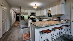 Una cocina moderna con armarios blancos, encimeras de granito, una isla central con taburetes de bar, electrodomésticos de acero inoxidable y una lámpara colgante.