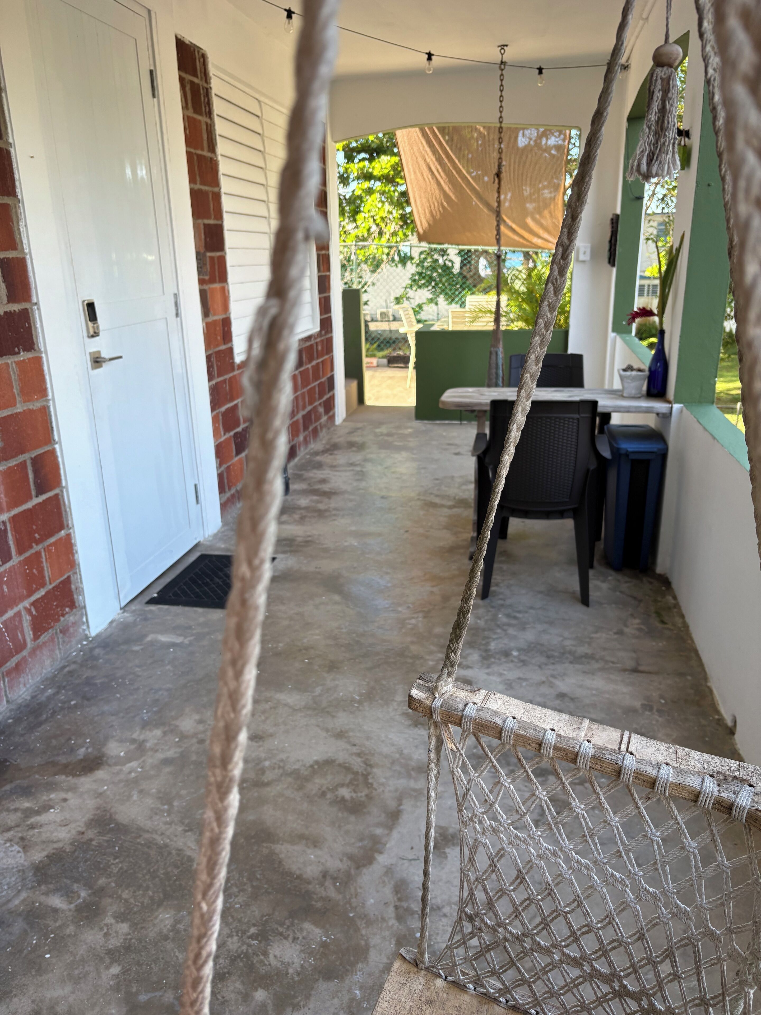 A covered outdoor area with a concrete floor, a white door, a brick wall, a table with chairs, and a hanging rope swing.