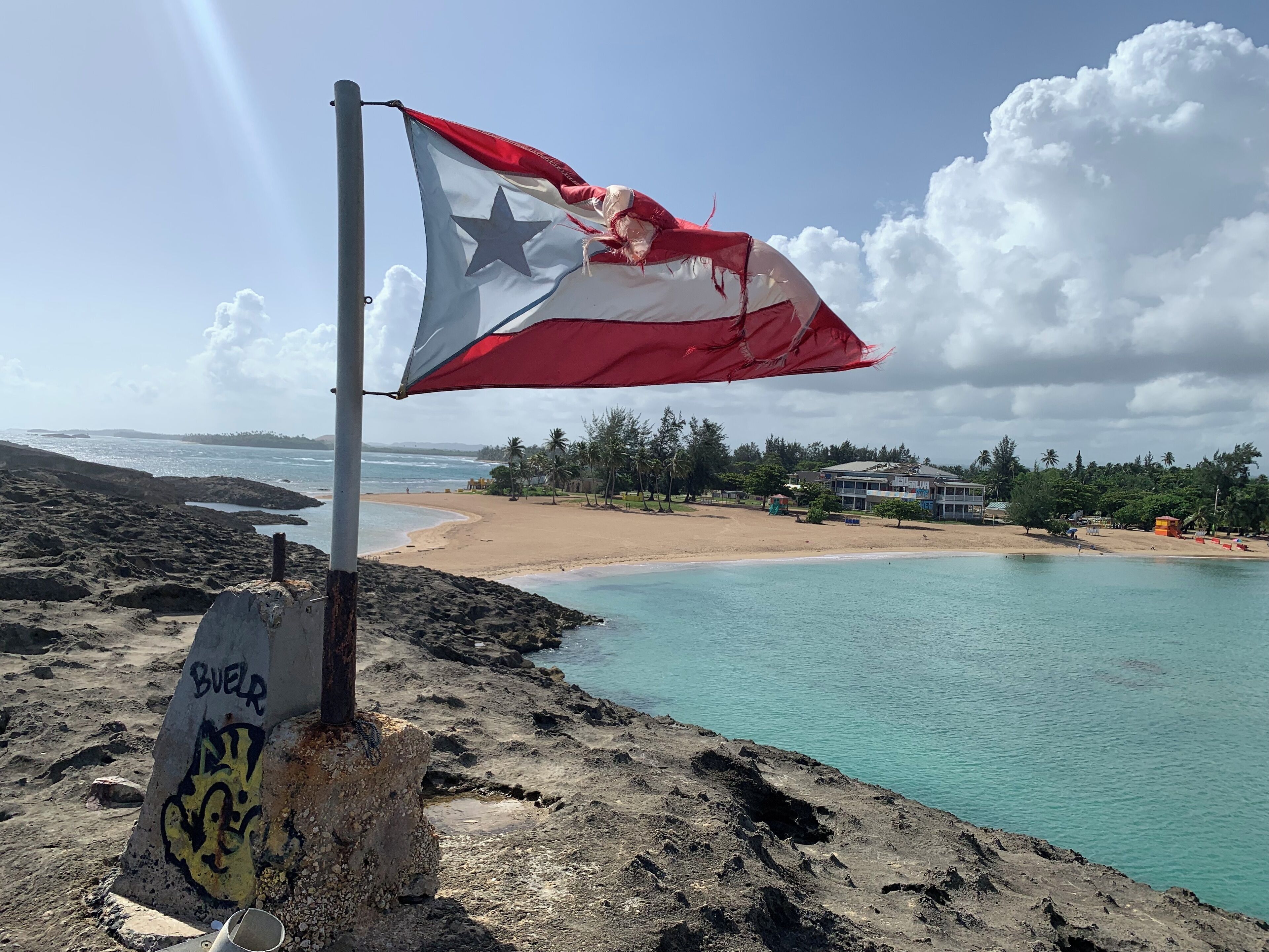 Un drapeau sur un mât, orné de graffitis, une plage et un bâtiment en arrière-plan.