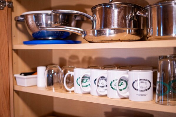 Una repisa de cocina con utensilios de acero inoxidable, una tabla de cortar azul y tazas blancas con logotipos verdes.