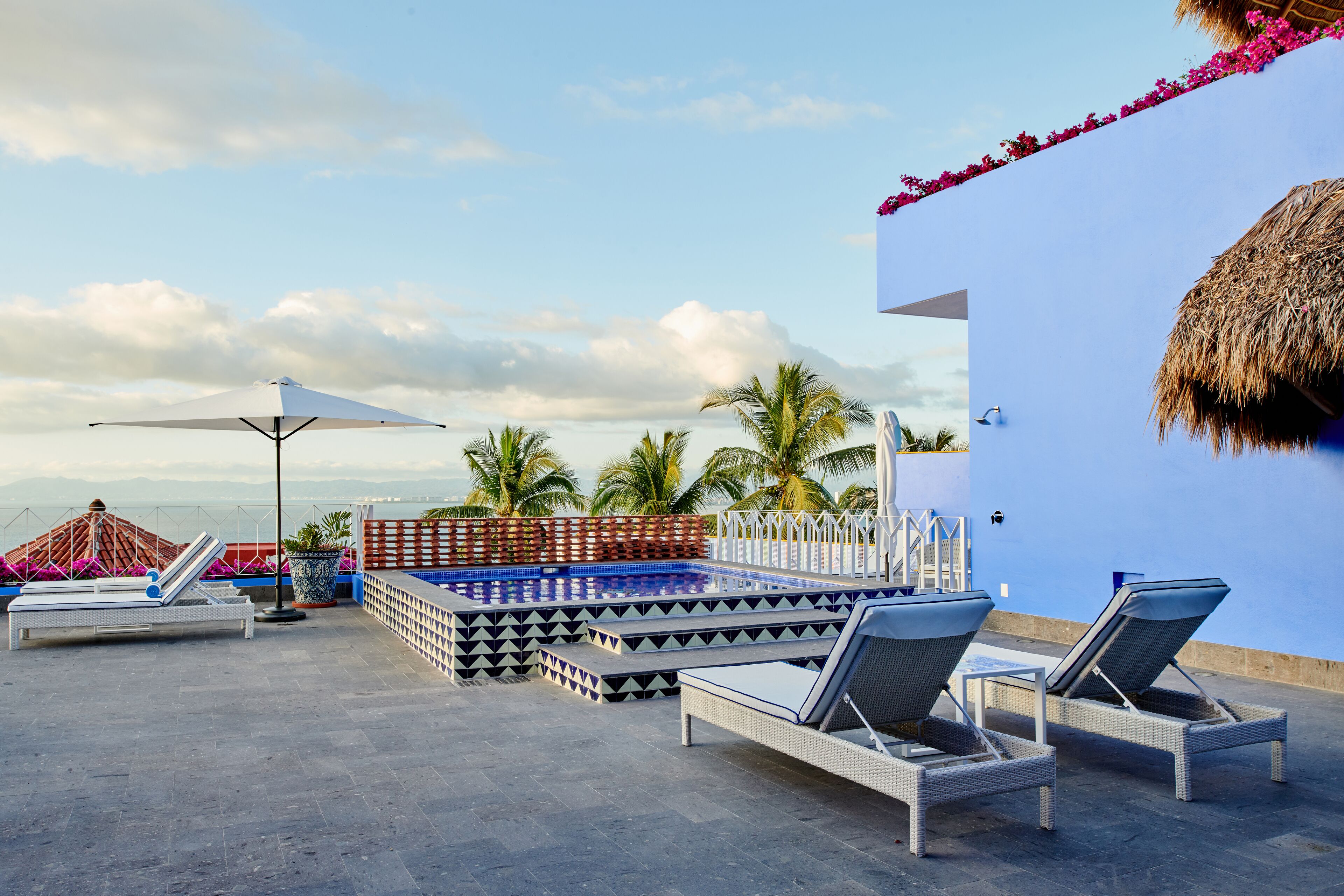 A rooftop pool area with lounge chairs, a pool, and a view of palm trees and mountains.