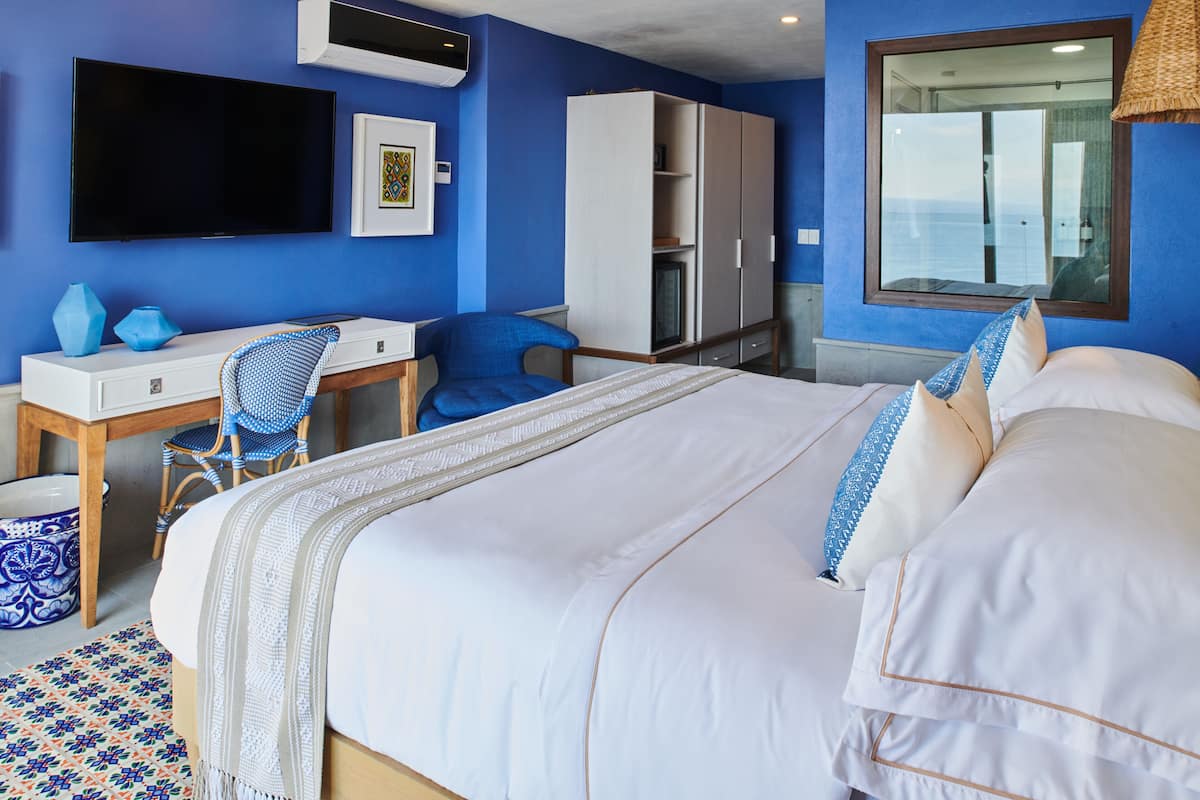 Habitación de hotel con cama, escritorio con silla, armario, televisión y un ventanal con vistas.
