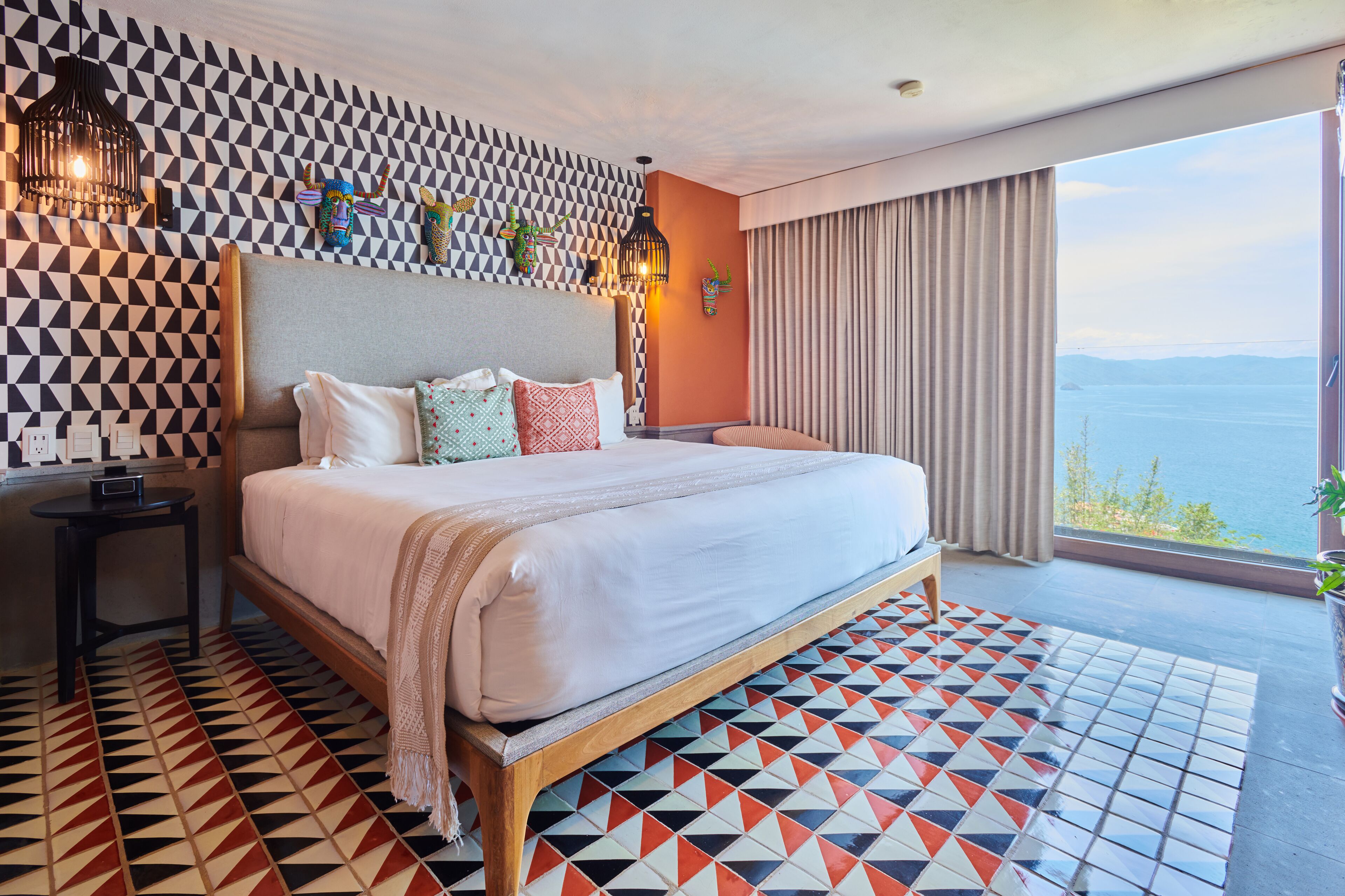 Quarto de hotel moderno com uma cama grande, piso com padrão geométrico e vista para o mar.