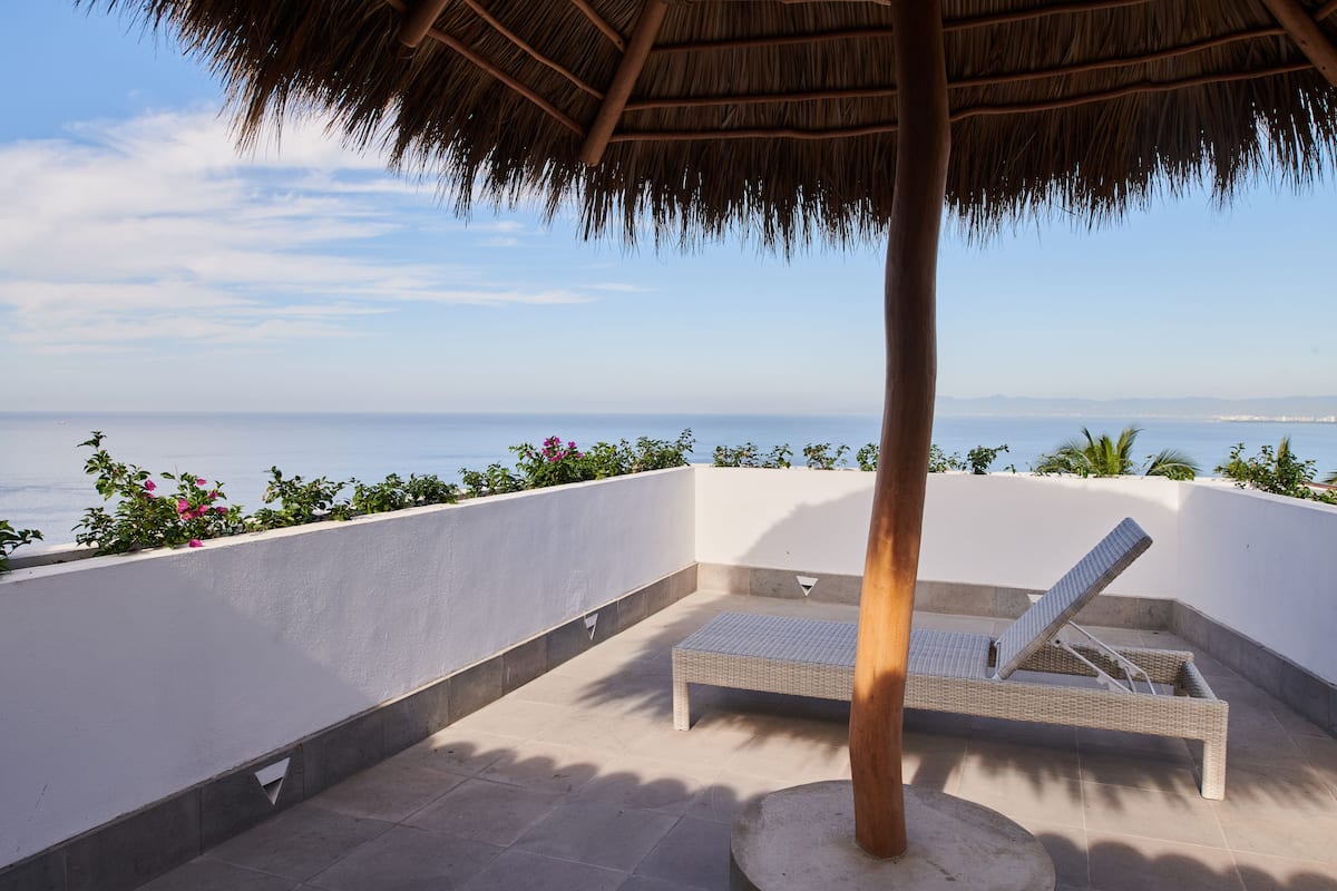 Un balcón con una silla lounge de mimbre, techo de paja y vista al mar.