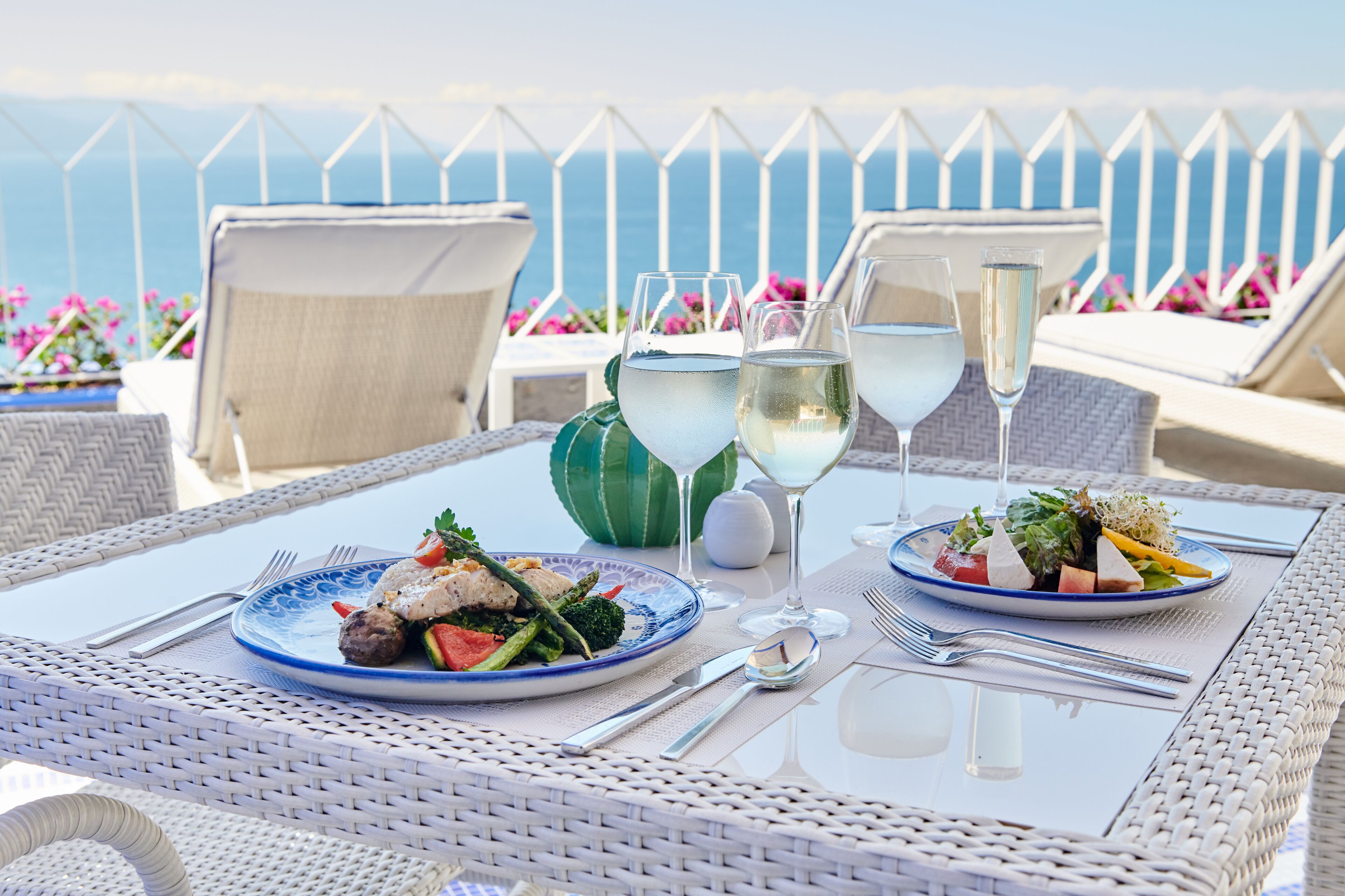 Ein Esstisch mit Tellern voller Essen und Weingläsern, auf einem Balkon mit Blick aufs Meer.