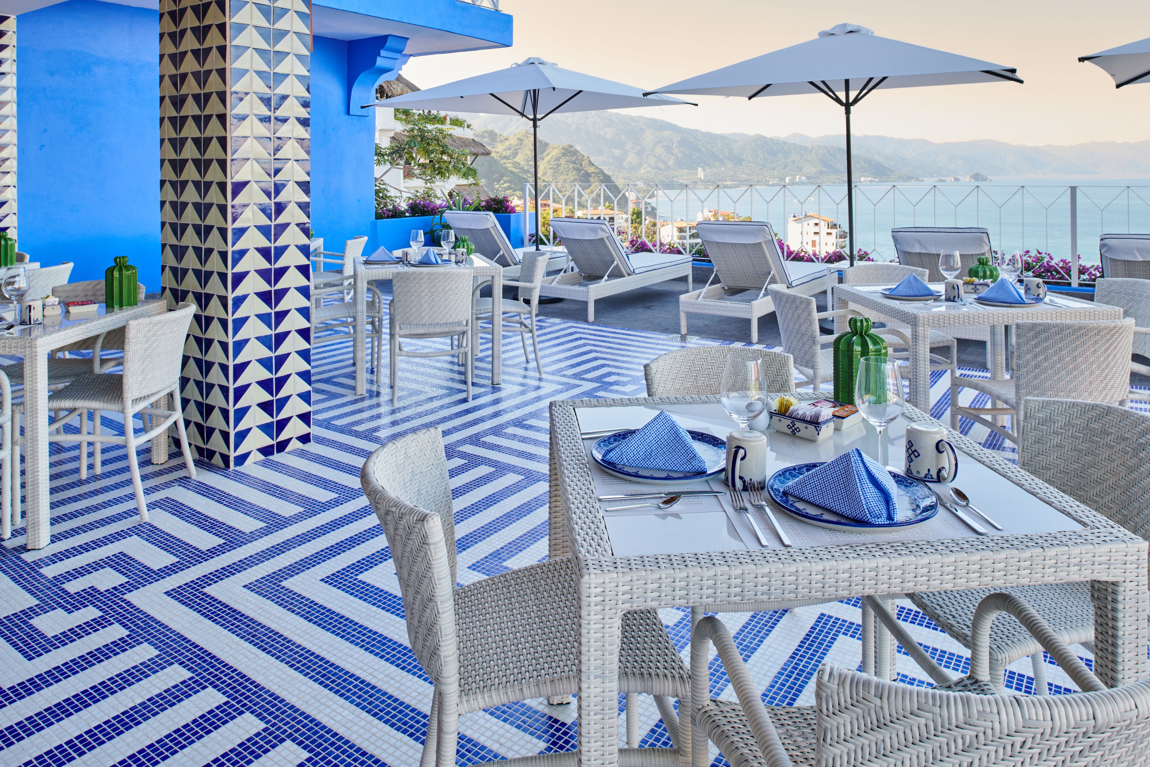 An outdoor dining area with white wicker furniture, blue and white patterned flooring, and umbrellas providing shade.