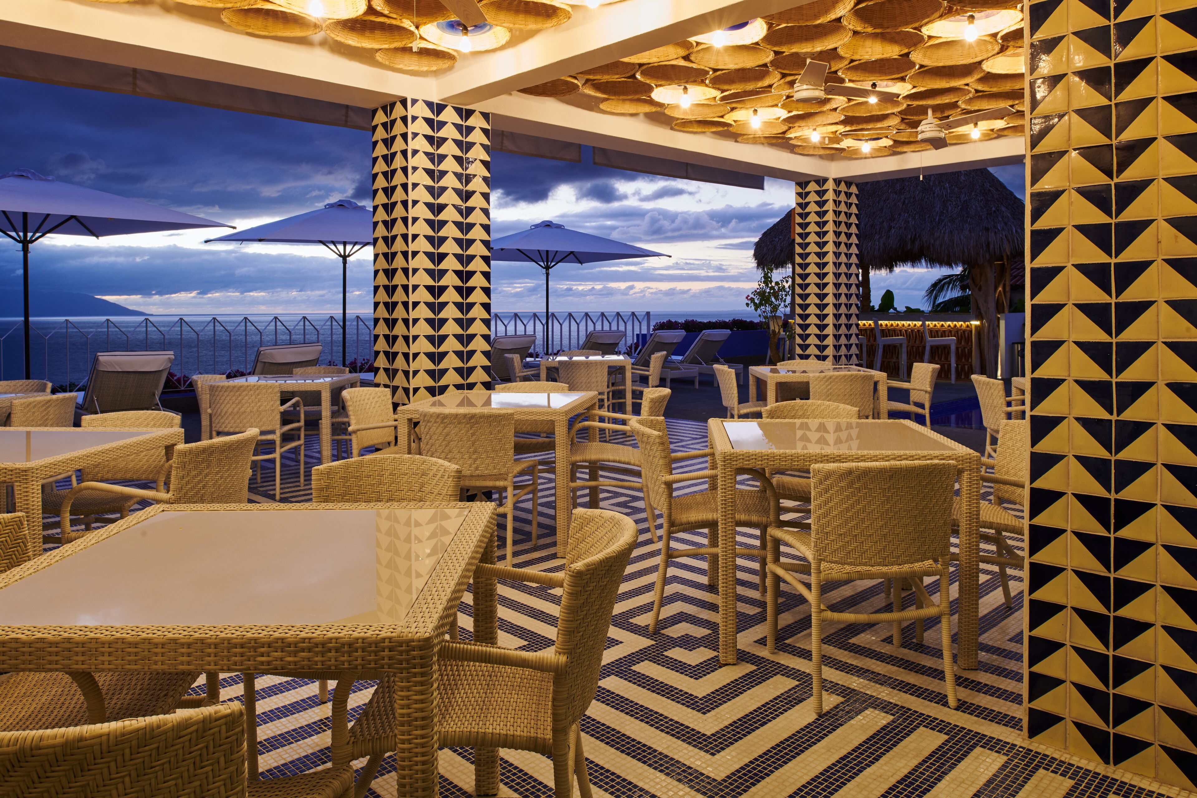 Un espacio de comedor al aire libre con mobiliario de mimbre y sombrillas, con vistas panorámicas.