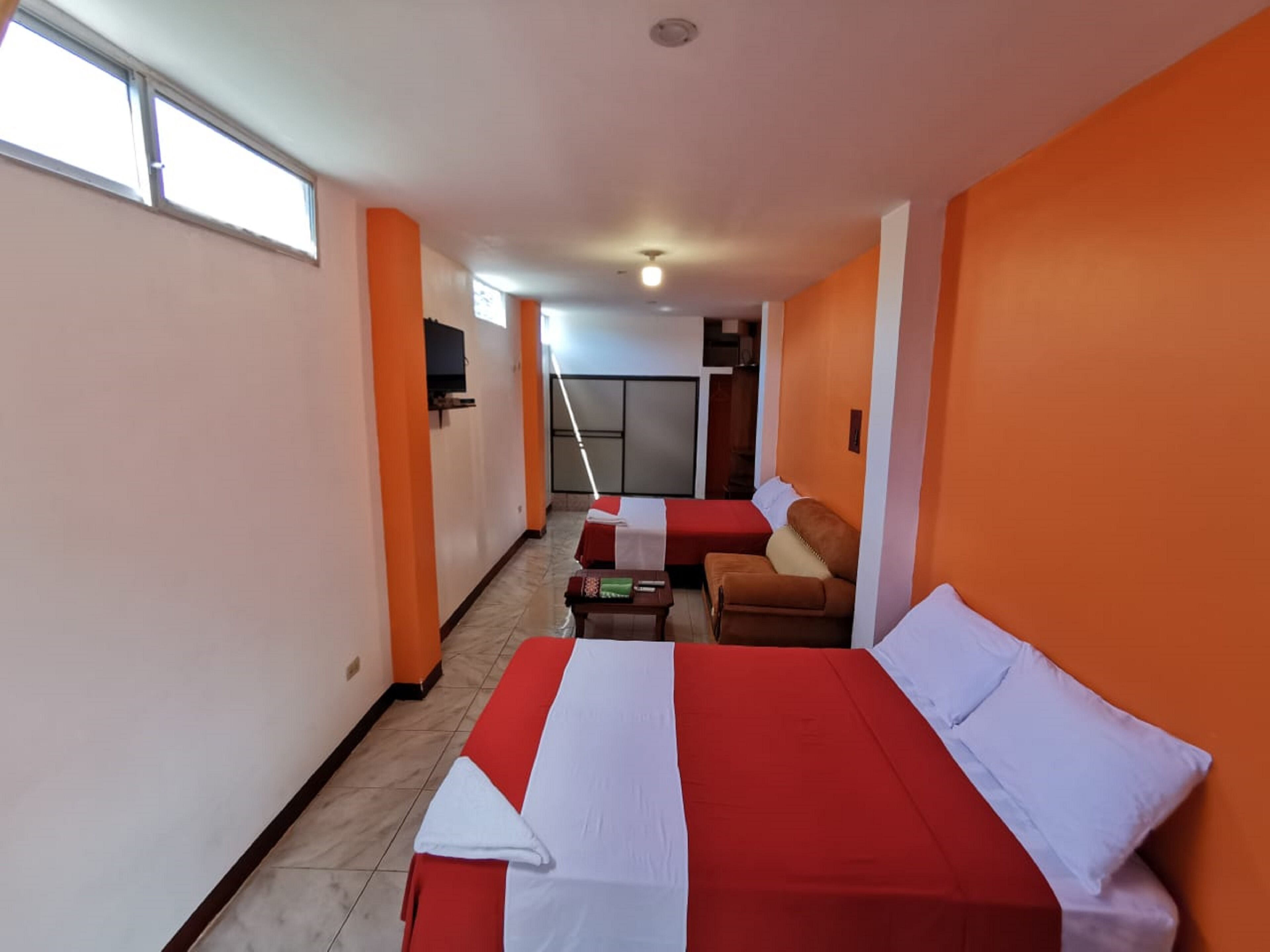 A hotel room with a large bed, a sofa, and a small table. There is a TV on the wall and a window providing natural light.