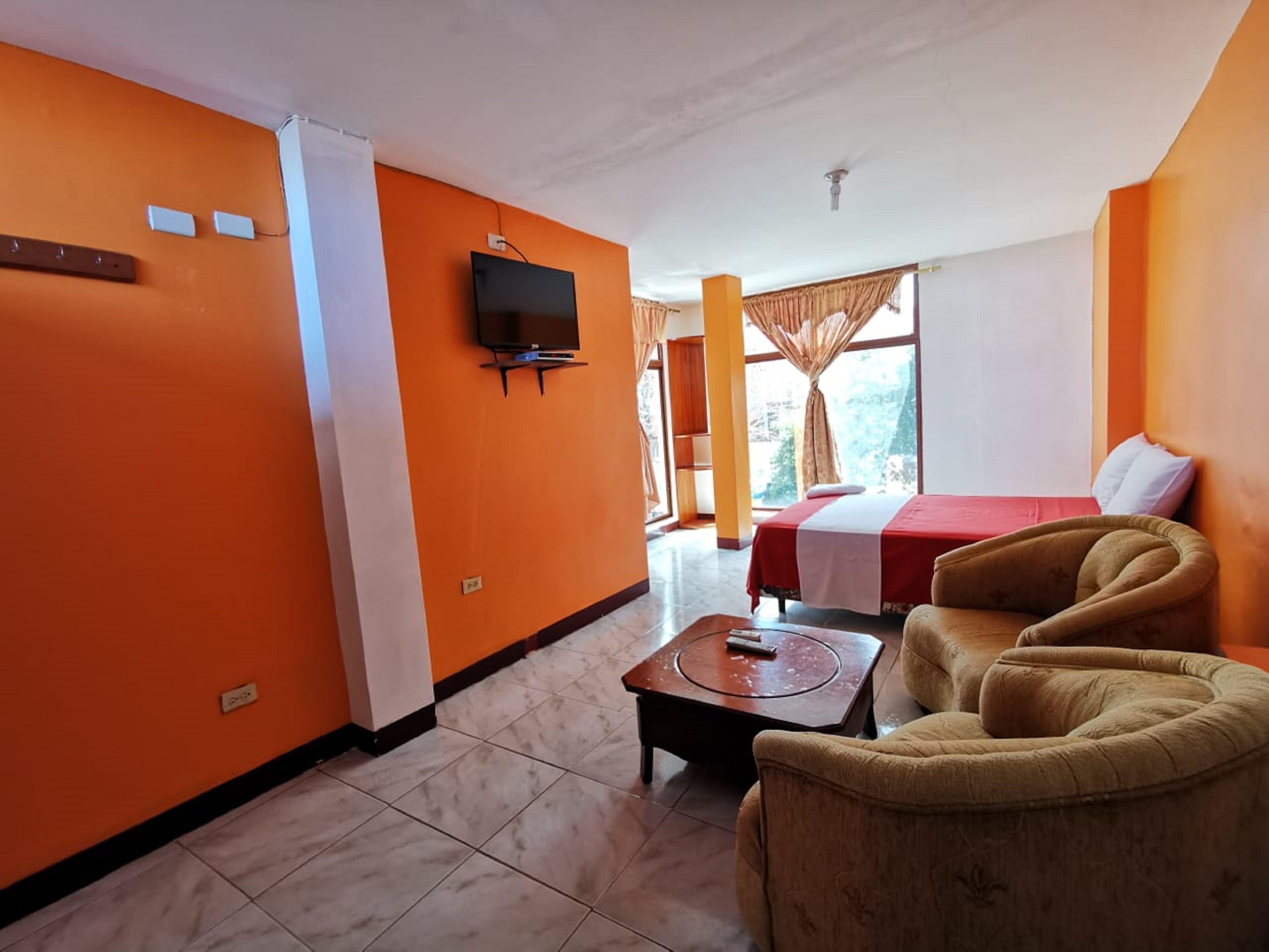 A hotel room with orange walls, a bed with a red and white bedspread, a television mounted on the wall, and a sofa.