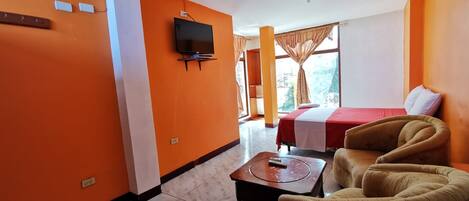 A hotel room with orange walls, a bed with a red and white bedspread, a television mounted on the wall, and a sofa.