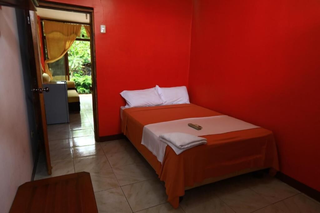 A small, red-walled room with a single bed, a wooden bench, and a door leading to a balcony with a view of greenery.