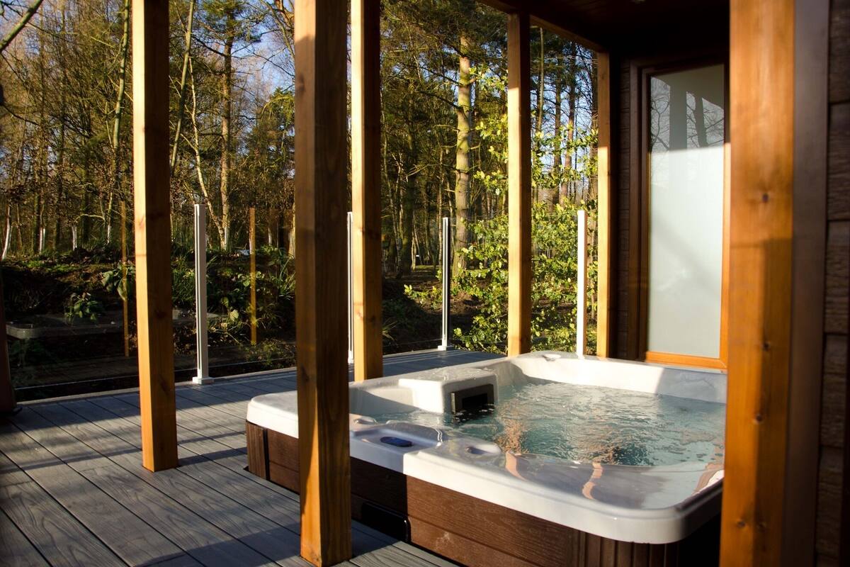 A wooden deck with a hot tub and a glass enclosure, surrounded by trees.
