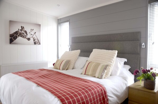 Un dormitorio con una cama, cabecera, radiador, ventana con persianas, un cuadro de jirafas en la pared y una mesita de noche con un florero.