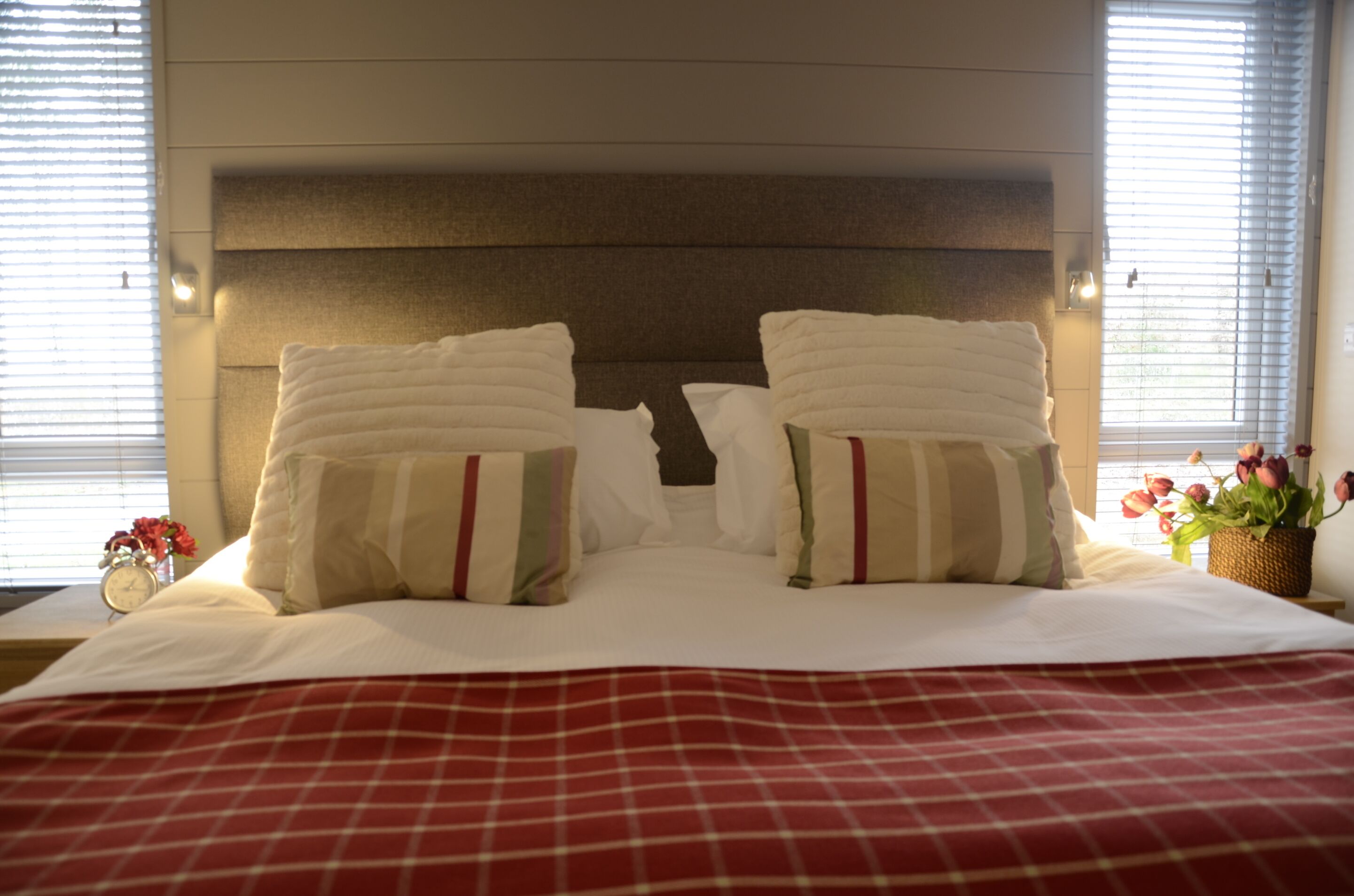 A neatly made bed with white linens and striped pillows, a red checkered bedspread, and a bedside table with a clock and flowers.