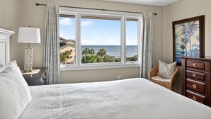 A bedroom with a large bed, a nightstand, a lamp, a chair, a dresser, and a window with a view of the ocean and palm trees. - PRIME Resort Location! Beachside, Ocean View, Pool Access! (Isle Of Palms)