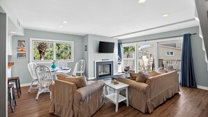 A spacious living room with a fireplace, beige sofas, a dining area with white chairs, and large windows overlooking a deck. - PRIME Resort Location! Beachside, Ocean View, Pool Access! (Isle Of Palms)