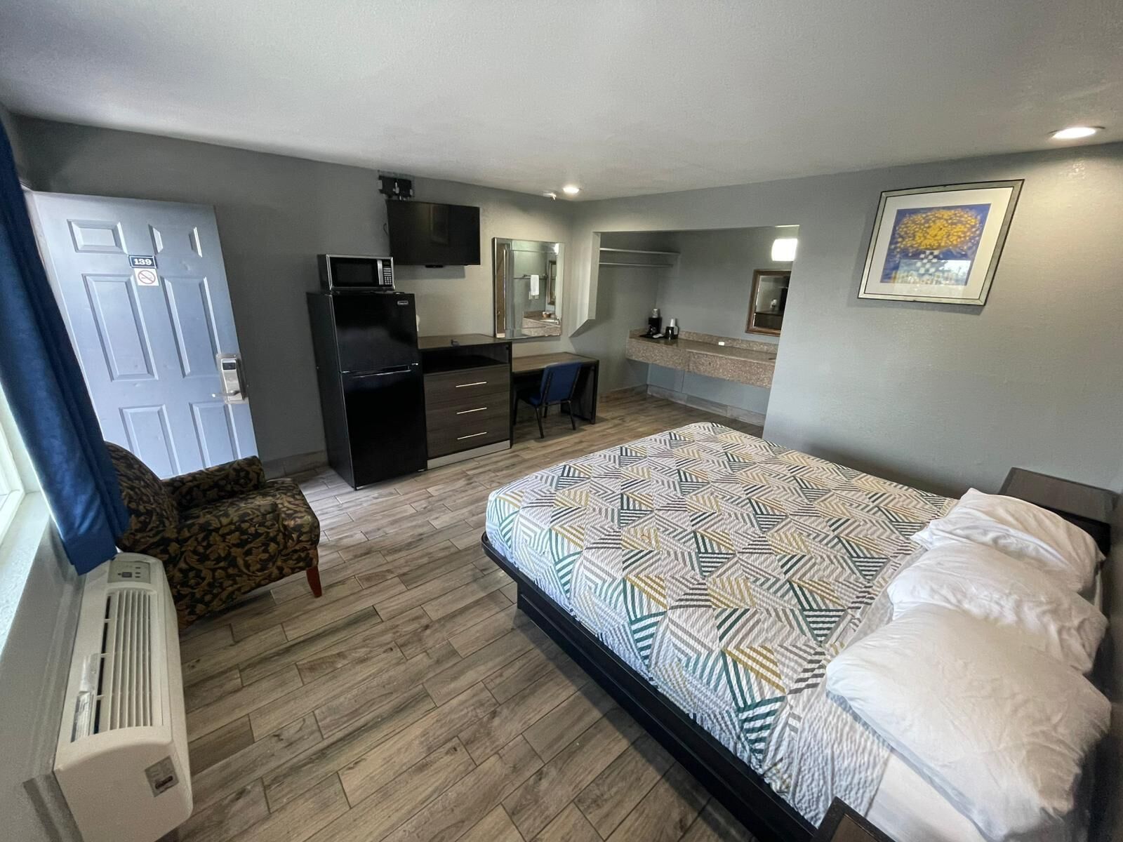 A compact bedroom with a bed, a desk, a chair, a TV, and a kitchenette.