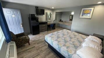 A compact bedroom with a bed, a desk, a chair, a TV, and a kitchenette.