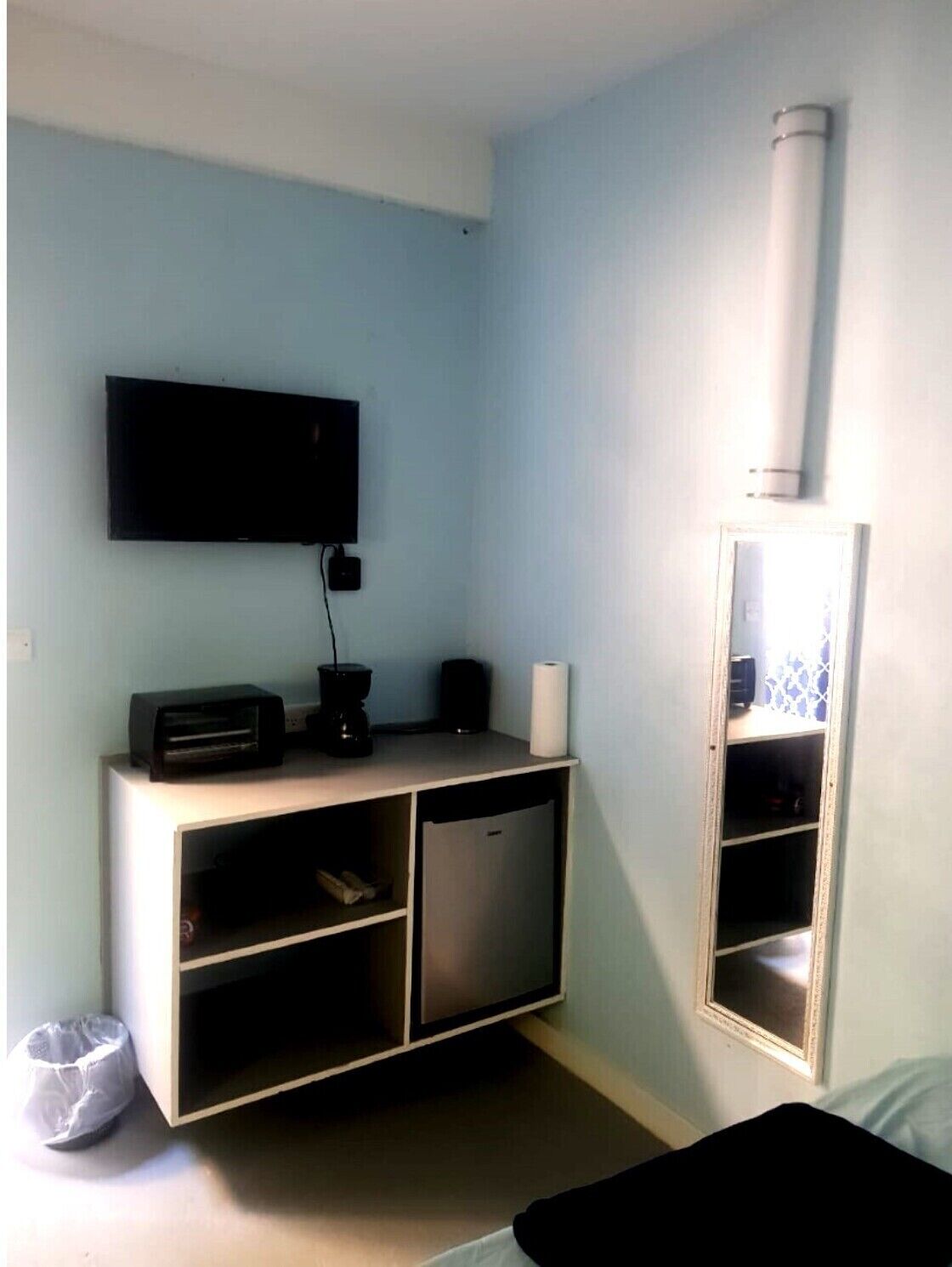 Una habitación con cama, televisión montada en la pared, kitchenette pequeño con cafetera y espejo en la pared.