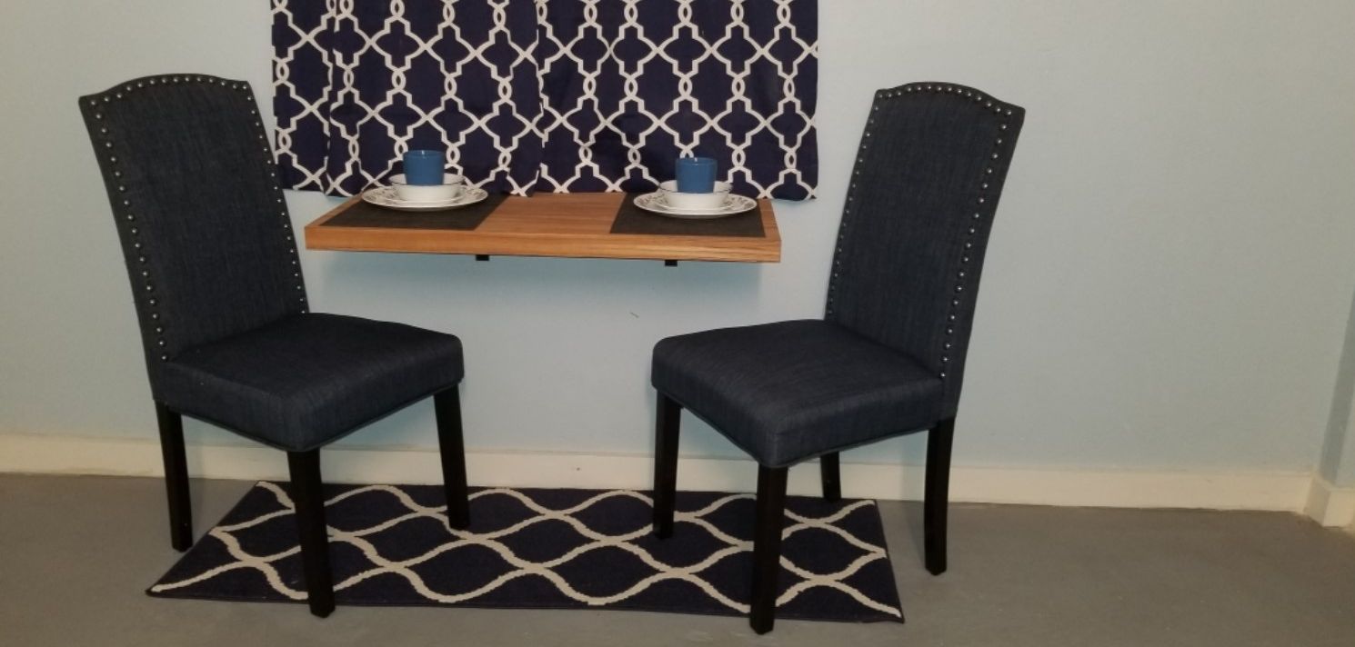 Dos sillones tapizados en azul oscuro con tachuelas de metal, un estante de madera con dos platos blancos y tazas azules, y una alfombra estampada en blanco y negro sobre una pared azul claro con cortina de diseño geométrico.