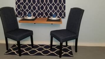 Dos sillones tapizados en azul oscuro con tachuelas de metal, un estante de madera con dos platos blancos y tazas azules, y una alfombra estampada en blanco y negro sobre una pared azul claro con cortina de diseño geométrico.