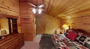 Een slaapkamer in een houten hutje, met een bed, een kapstok en een plafondventilator.
