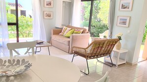 A dining area with a white table and chairs, a wicker chair, a sofa with cushions, and a view of greenery outside.