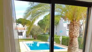 A room with a view of a swimming pool, a palm tree, and a window with blinds.