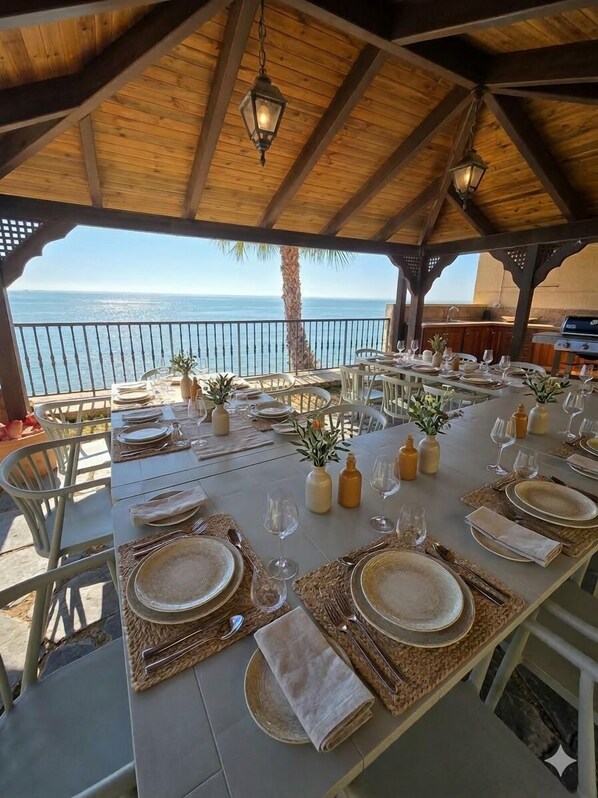 An outdoor dining area with a view of the sea, wooden beams, and hanging lanterns.