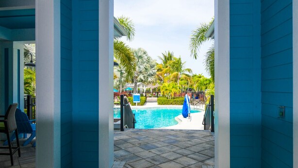 A pool area with a clear view of the pool, surrounded by palm trees and a blue building facade. - Changes in Latitude! Gulf Views and Pool! (Bradenton)