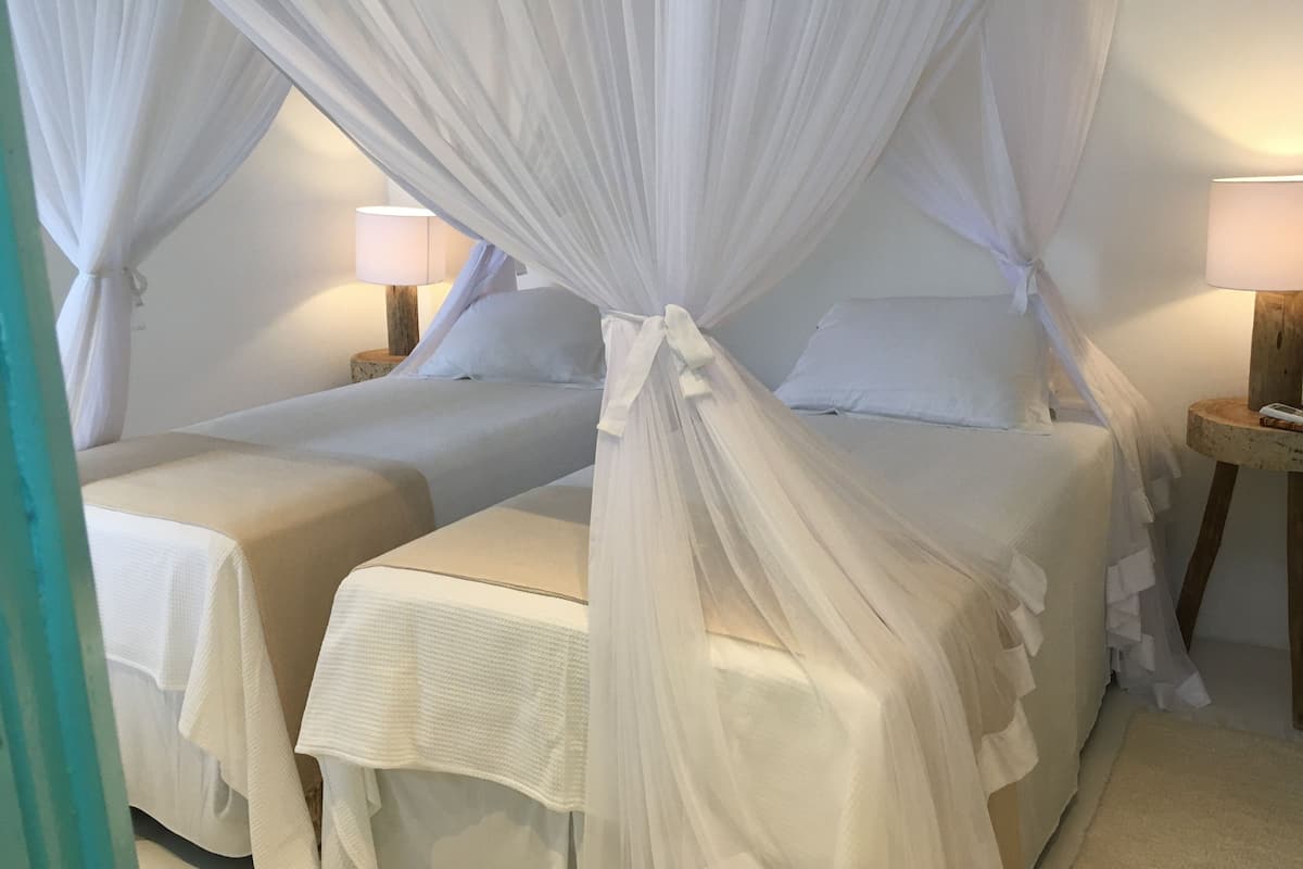 Cama de casal com dossel, duas mesas de cabeceira e um criado-mudo com luminária.
