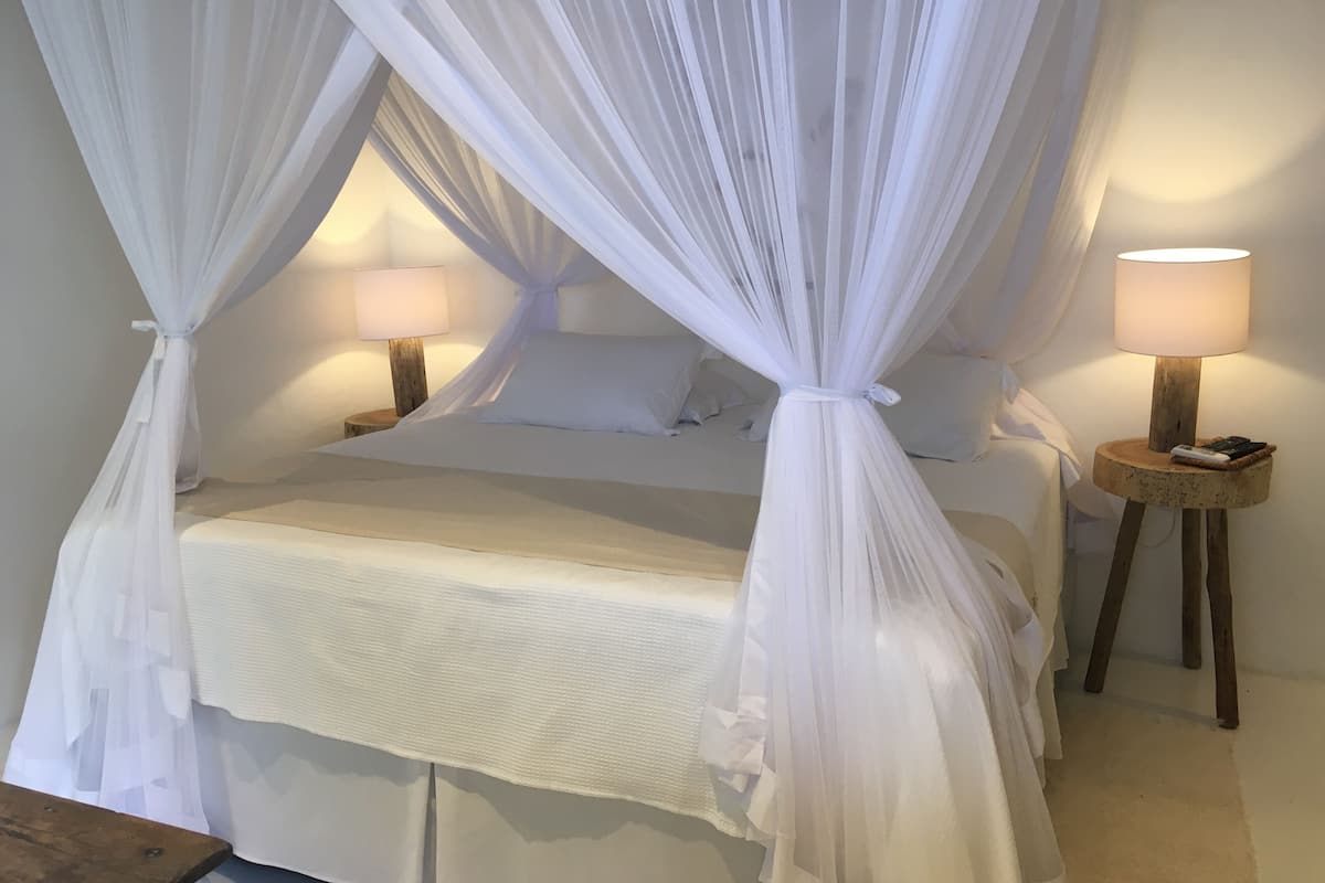 Quarto com cama de dossel, duas mesas de cabeceira com luminárias e uma mesinha de cabeceira redonda de madeira.