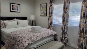 Habitación de hotel con cama, lámpara de noche, banqueta, un cuadro en la pared y ventana con cortinas florales.