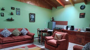 Ein Wohnzimmer mit einem roten Sofa, einem roten Sessel und einem Essbereich mit Tisch und Stühlen. An den Wänden befinden sich Regale mit Pflanzen und gerahmten Bildern.