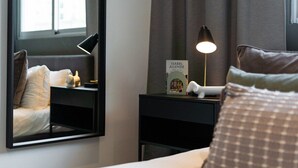 Una habitación de hotel con cama, mesita de noche con lámpara, libro enmarcado y un espejo que refleja el cuarto.