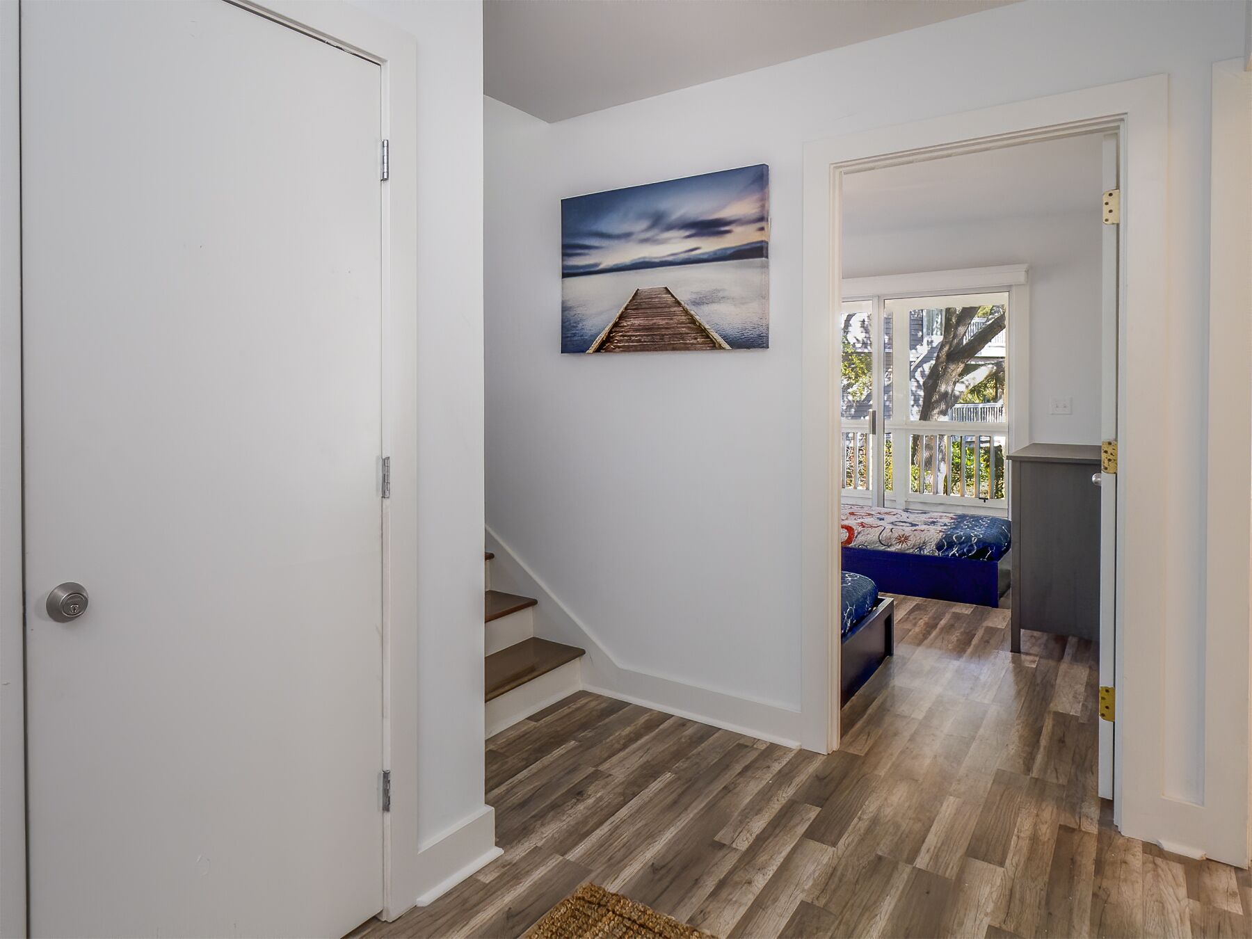 A hallway with a wooden floor, a white door, and a staircase leading to a room with a bed and a window.