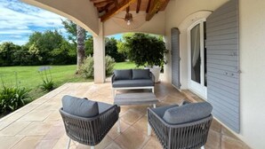 Un patio cubierto con techo de madera, un sofá gris, dos sillas de mimbre y una planta en maceta.