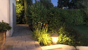 Un sendero conduce a un edificio de fachada blanca, rodeado de exuberante vegetación y un jardín bien iluminado.