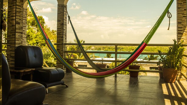 A hammock on a balcony with a view of the ocean and a pool. There are chairs and potted plants on the balcony.