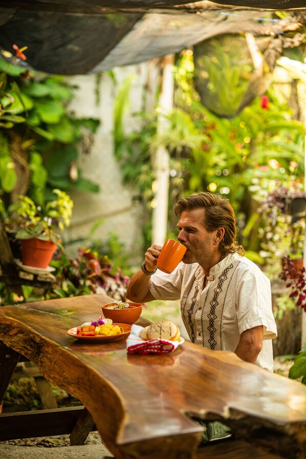 Una persona sentada a una mesa de madera al aire libre, con diversas plantas y una maceta de fondo.