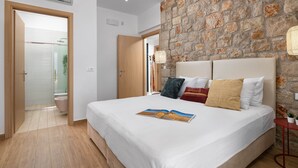 Un dormitorio con pared de piedra, cama con muchos almohadones, mesita de noche con lámpara y puerta que da a un baño.