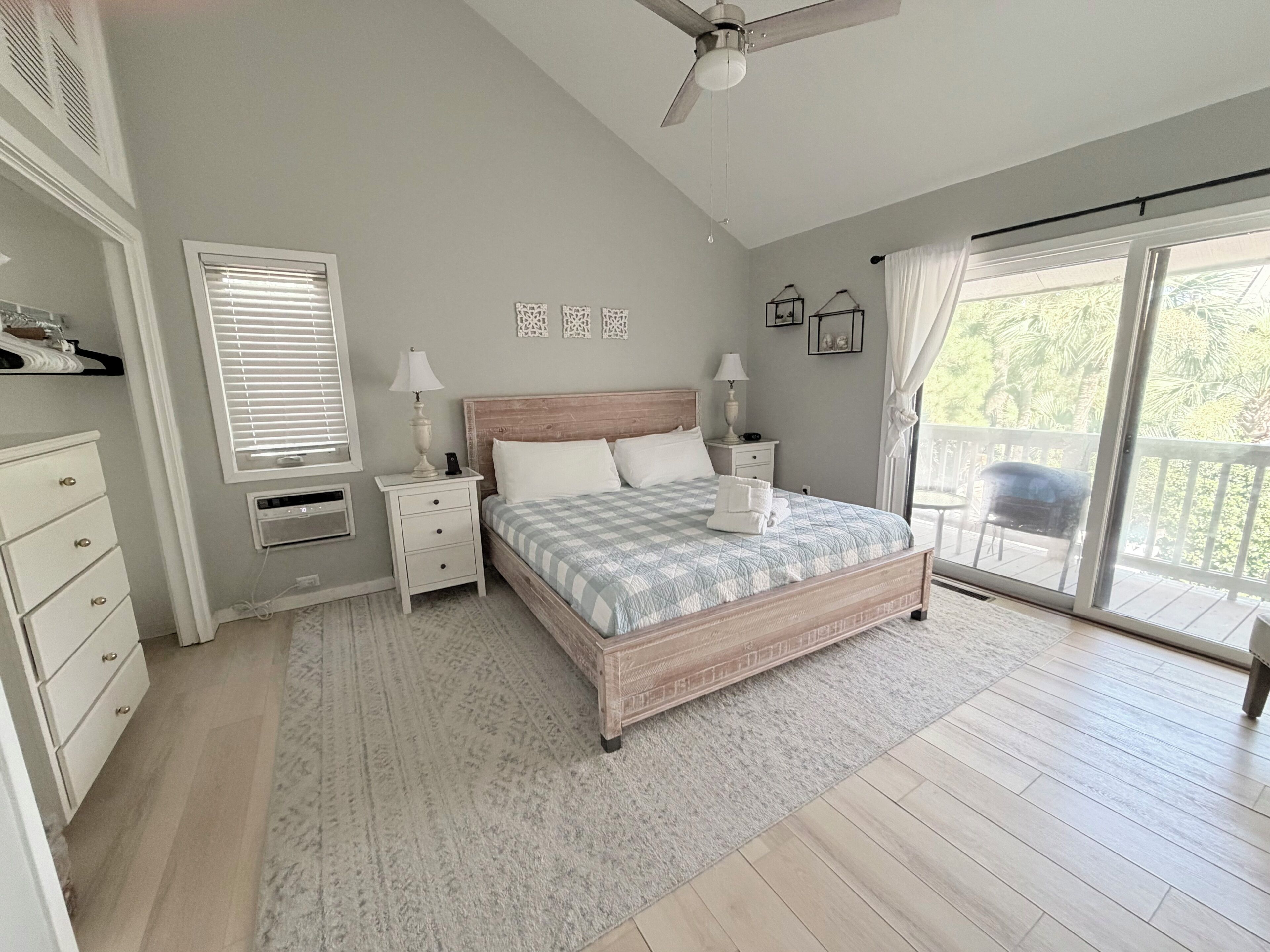 A bedroom with a bed, bedside tables, a ceiling fan, a window with blinds, a sliding glass door, and a view of greenery outside.
