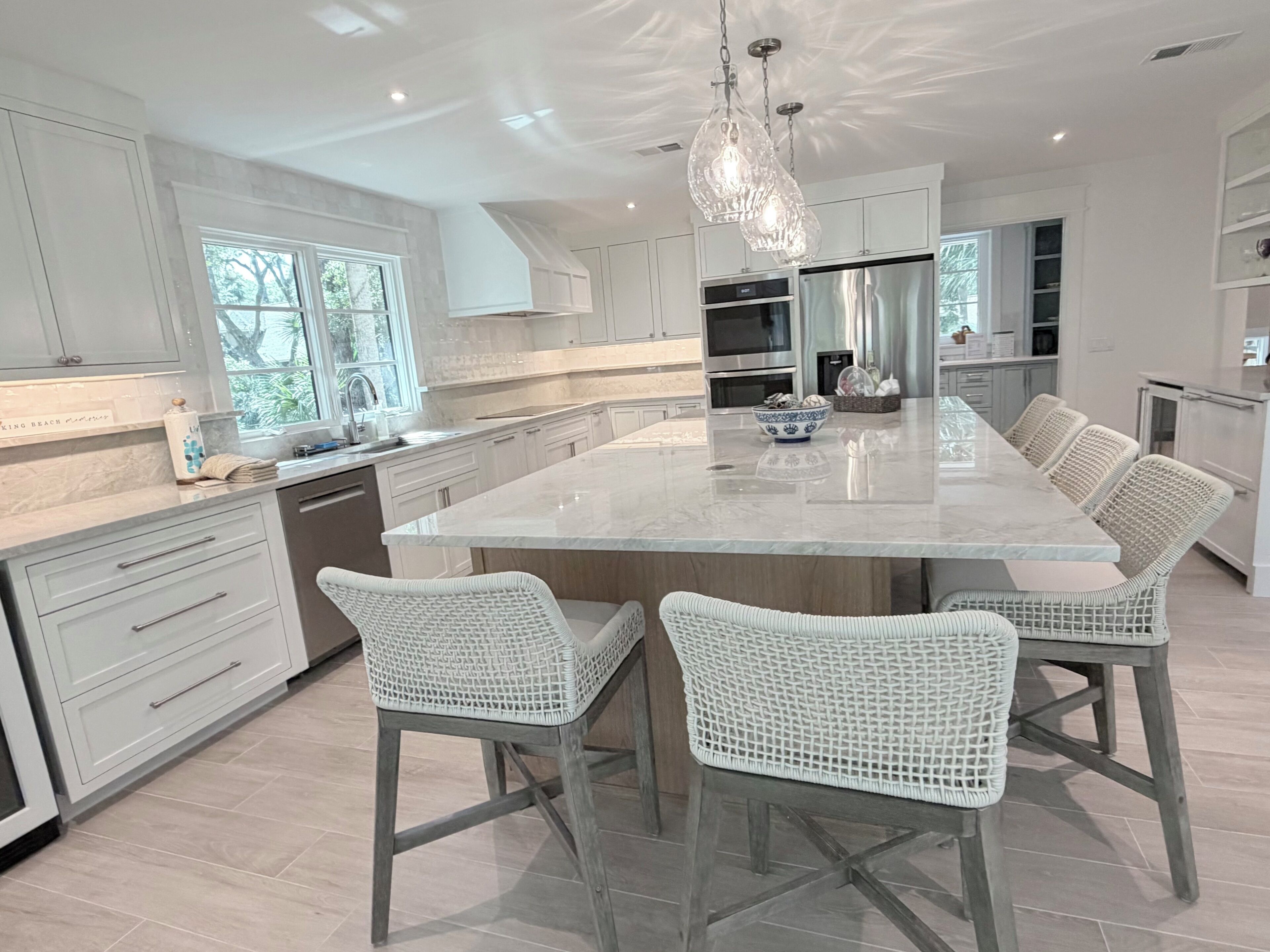 A modern kitchen with white cabinetry, a large island, wicker chairs, and pendant lighting.
