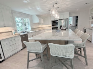 Cocina moderna con armarios blancos, una isla grande, sillas de mimbre e iluminación colgante.