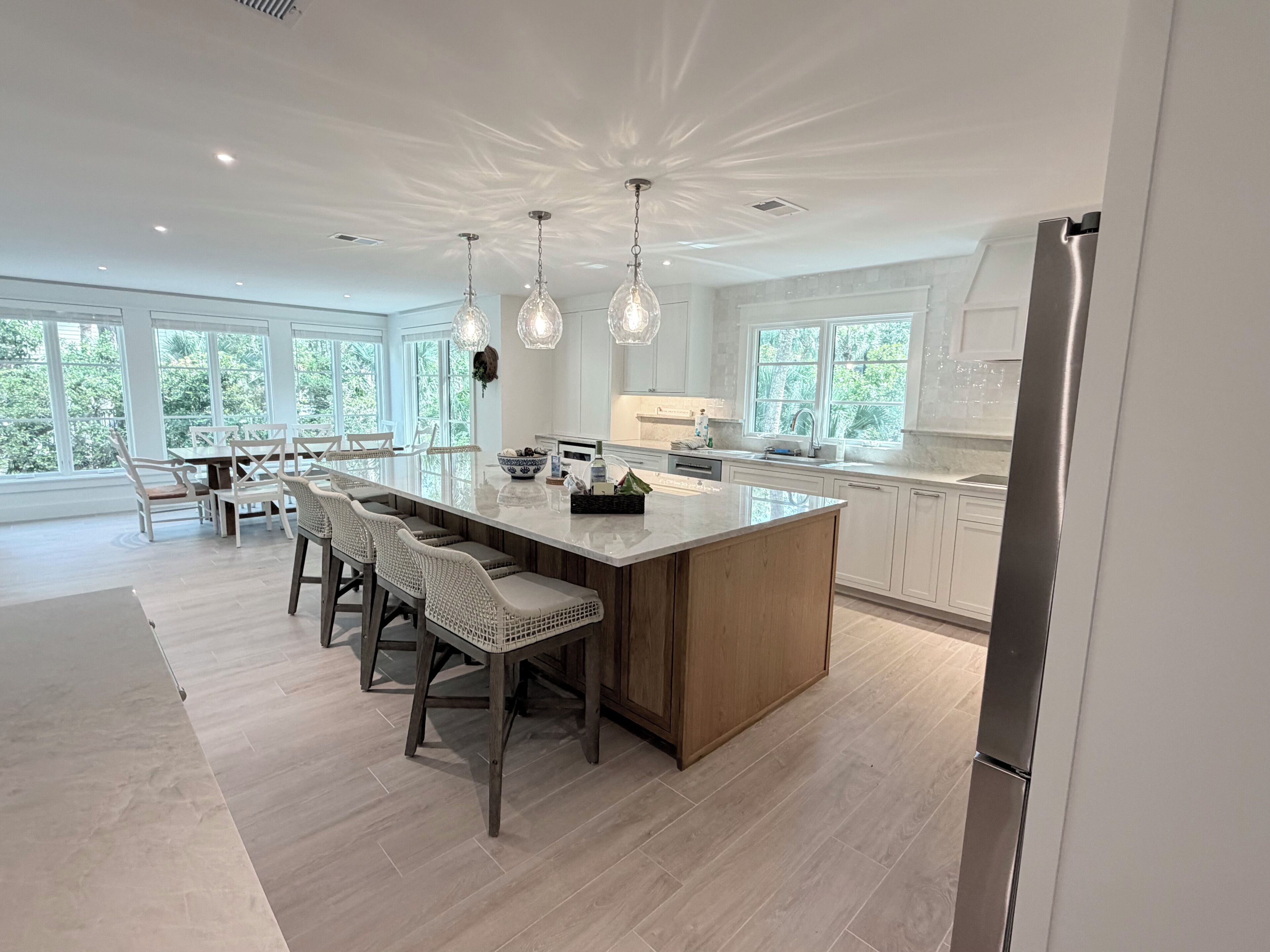 A modern kitchen with a large island, pendant lights, and a view of greenery outside.