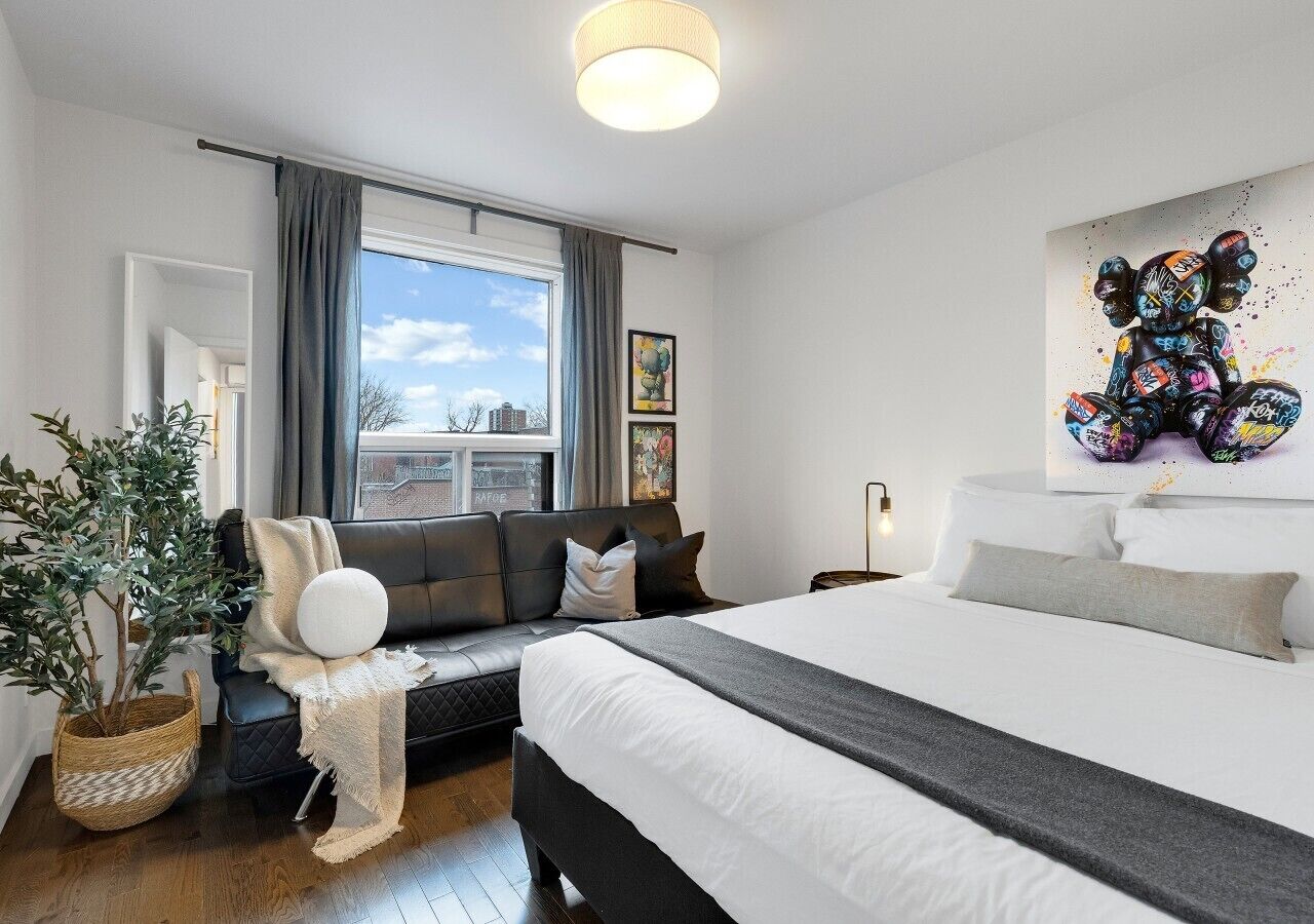 A modern bedroom with a large bed, a sofa, a potted plant, and a city view.