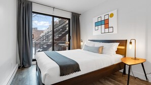 Ein modernes Hotelzimmer mit einem großen Bett, Blick auf die Stadt durch ein Fenster und einem minimalistischen Kunstwerk an der Wand.
