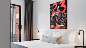 Hotelkamer met een groot bed, twee nachtkastjes, een poster aan de muur en uitzicht op een bakstenen gebouw door een raam.