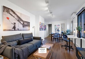 A modern living room with a grey sofa, a dining area with a table and chairs, and a kitchen area with a counter and a plant.