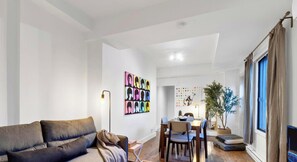 Uma sala de estar com sofá, mesa de jantar com cadeiras, uma planta e uma parede com obras de arte coloridas.